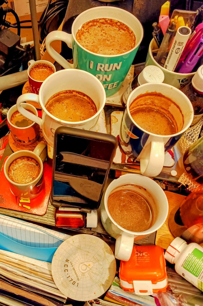 Top-down view of 3 full espresso cups and and 4 mostly-full mugs of coffee.- o a cluttered table.